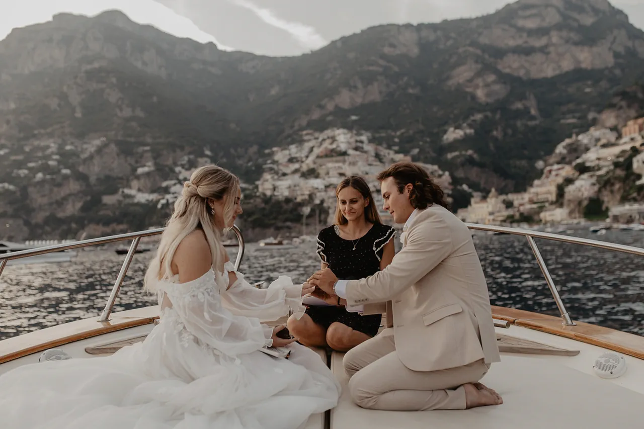 Wedding in Positano. A couple is sitting on the deck of a boat having a wedding ceremony, with a woman officiating. The bride is wearing a white dress, and the groom is in a light suit. They're surrounded by scenic mountains and a coastal town in the background.