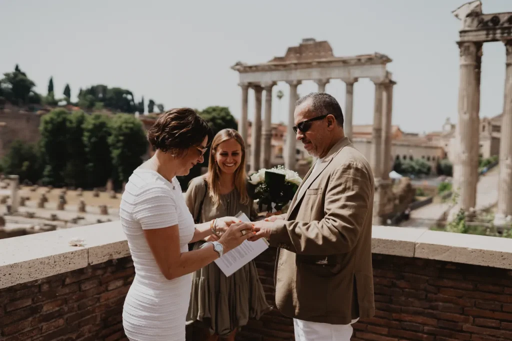 A couple is exchanging vows outdoors near ancient ruins. The bride in a white dress and the groom in a tan suit are smiling. A woman stands behind them holding a bouquet. Columns and a ruin are visible in the background.