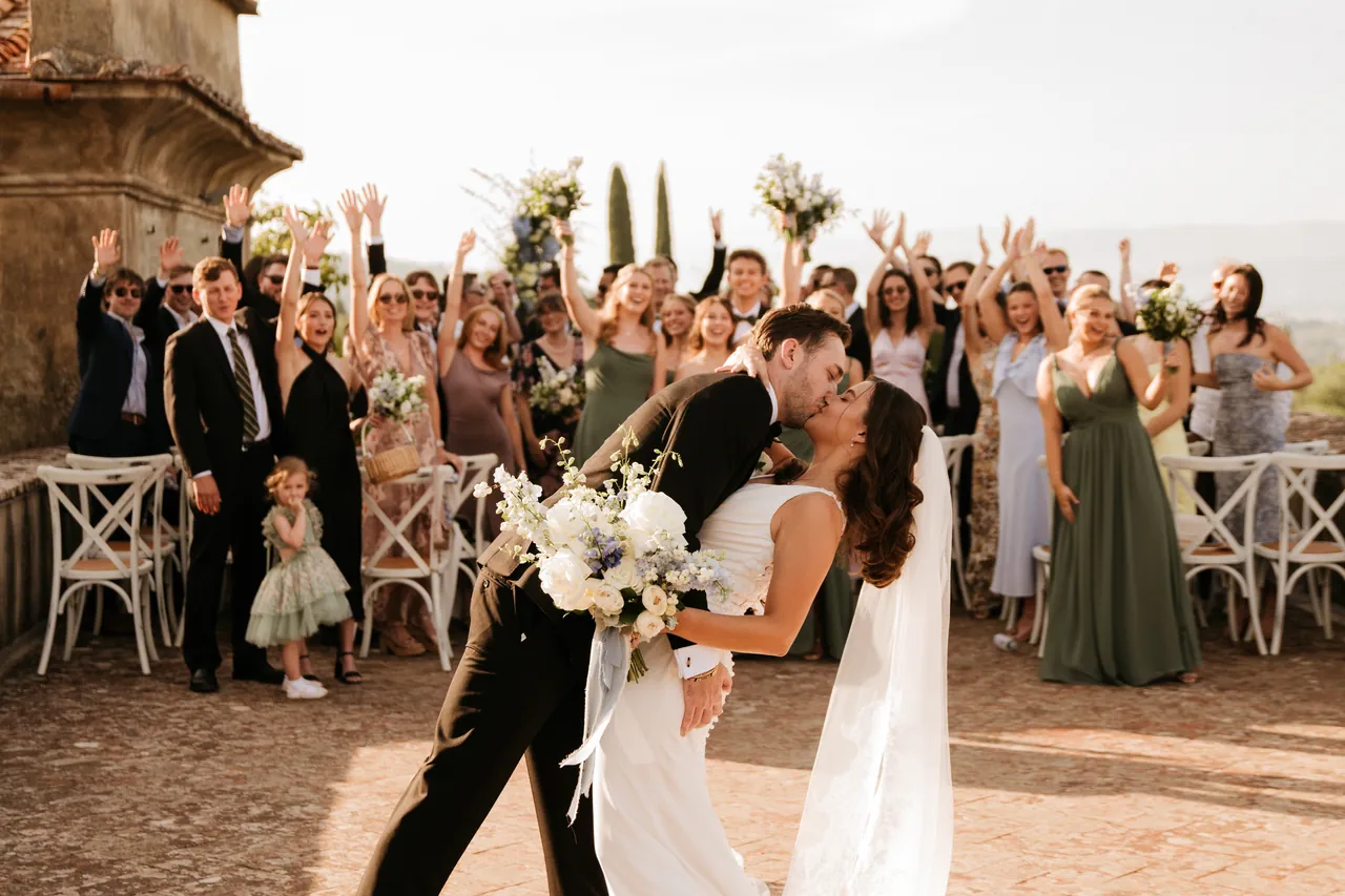 Chianti wedding. A newlywed couple shares a kiss as the groom dips the bride, who holds a bouquet. They're surrounded by cheering friends and family in formal attire, set against the scenic backdrop of a wedding in Chianti under a clear sky.